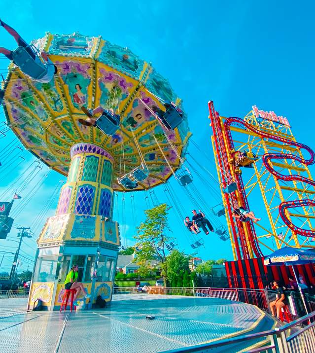 A vibrant Long Island amusement park scene featuring a spinning swing ride and a colorful roller coaster against a clear blue sky.