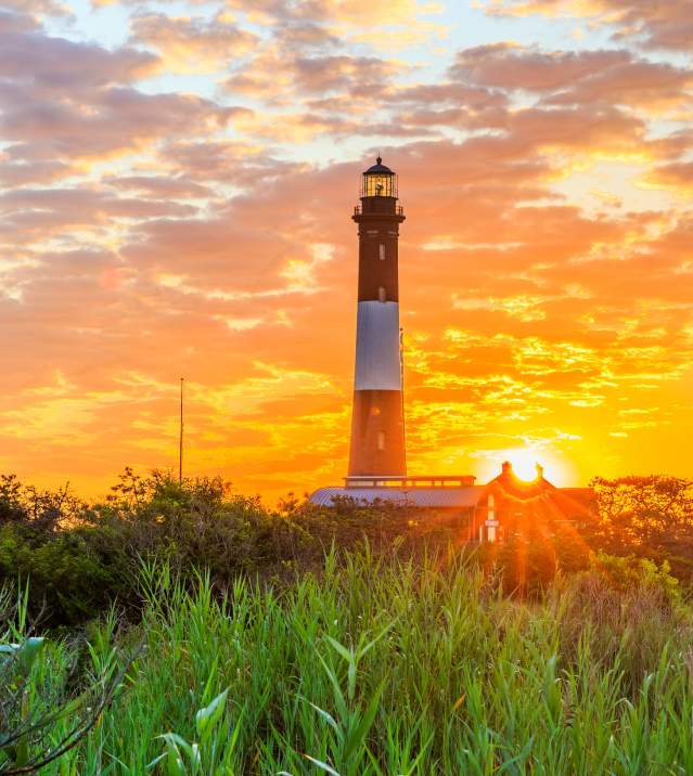 Tall lighthouse rising above coastal grasses on Long Island at sunset, with a glowing orange sky, scattered clouds, the sun near the horizon, and warm light illuminating the surrounding landscape.