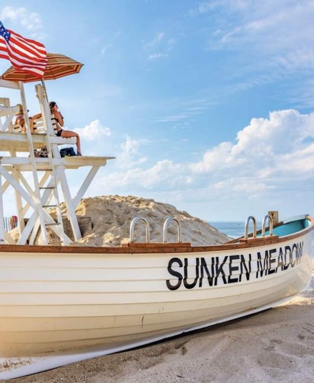 Wooden lifeguard chair and small boat labeled “SUNKEN MEADOW” resting on sandy Long Island beach, with an American flag mounted on the structure, dunes and beach fencing behind, and calm ocean water under a bright blue sky.