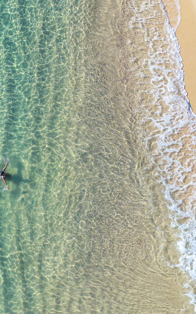 A person swimming in clear turquoise waters near a sandy beach, viewed from above.