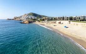 An aerial, high-angle view of the crystal-clear, turquoise waters and golden sands of Chileno Bay in Los Cabos. The swimmable beach is lined with modern luxury resorts and framed by rugged desert hills under a bright, clear blue sky.