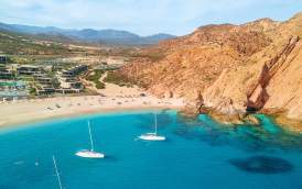 An aerial view of the vibrant turquoise waters of Santa Maria Bay in Los Cabos, featuring three white sailboats anchored in the calm, horseshoe-shaped cove. The golden sand beach is framed by dramatic reddish-brown cliffs and the modern architecture of a luxury resort under a bright blue sky with wispy white clouds.