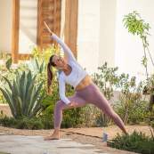 A woman with a ponytail wearing a white long-sleeve crop top and lavender leggings holds an extended side angle yoga pose. She is practicing on a mat in an outdoor garden area featuring stone tiles, gravel landscaping, and a large agave plant.