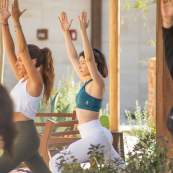 A side-profile close-up of women in a yoga class holding a high lunge pose with their arms raised straight up. The central woman wears a teal sports bra and white leggings with her eyes closed in focus amidst a sunny outdoor setting.