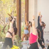 An outdoor yoga class at Anima Village shows participants in various activewear stretching their arms overhead and leaning to the side. The class is held on a stone patio with large wooden pillars next to a white wall featuring the 'ANIMA VILLAGE' logo.