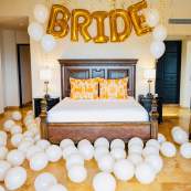 A luxury villa bedroom decorated for a bachelorette party with gold "BRIDE" balloons and a floor covered in white balloons.