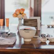 A curated skincare workshop setup on a polished wooden table featuring Natura Bissé products. In the center, a branded white ceramic bowl holds a treatment packet, surrounded by a folded white towel with a headband, applicator brushes, and orange skincare bottles. A small vase of peach roses and a tabletop mirror sit in front of a blurred coastal view background.