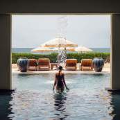 A woman in a dark swimsuit stands under a cascading water feature in an indoor-outdoor spa pool. Framed by architectural pillars, the view opens onto a resort sun deck equipped with lounge chairs, large cream-colored umbrellas, decorative ceramic urns, and the ocean horizon in the distance.
