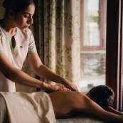 A female spa therapist in a light-colored uniform gives a back massage to a client lying face down on a massage table. The serene treatment room features patterned curtains and a glimpse of an outdoor plunge pool through an open doorway in the background.