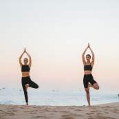 Two women in black athletic wear practice the tree yoga pose on a sandy beach at dusk. They stand balanced on one leg with their hands pressed together overhead, against a backdrop of a calm ocean and a soft, pastel sky.