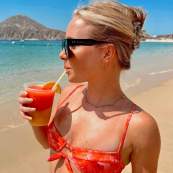 A woman in a floral orange bikini and designer sunglasses enjoys a vibrant, layered frozen tropical cocktail on the golden sands of Medano Beach. The turquoise waters of the Sea of Cortez and the blurred silhouette of the iconic Land's End rocks.
