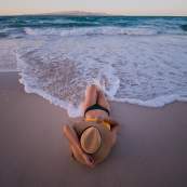 A high-angle, peaceful view of a traveler lying on the soft sand of a Los Cabos beach, with a wide straw hat covering her face. Gentle, foamy white waves from the turquoise Sea of Cortez wash up toward her feet during the soft light of dusk, with the distant silhouette of Baja’s rugged mountains on the horizon.