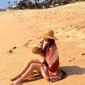 A woman in a wide-brimmed straw hat and a flowing orange tie-dye cover-up sits on the vast golden sands of a Los Cabos beach, enjoying a fresh coconut drink through a straw. The background features a luxury beachfront resort with traditional thatched palapa roofs, palm trees, and white sun umbrellas under a clear, bright blue sky.