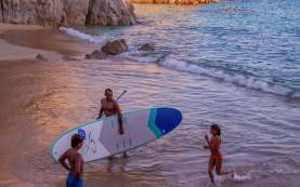 A family enjoys the soft golden hour light at a secluded beach in Los Cabos. A father carries a blue and white stand-up paddleboard through the shallow turquoise surf while his young daughter splashes in the waves and his son watches from the shore. The small cove is framed by a massive, sun-drenched granite cliff, with the distant skyline of Cabo San Lucas visible across the bay.