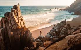 A cinematic view of a lone traveler standing atop a rock at a secluded beach in Los Cabos, dwarfed by massive, rugged granite formations. The late afternoon sun casts a golden glow over the breaking white waves and the sandy shore, perfectly capturing the dramatic "desert-meets-sea" landscape of the Baja peninsula.