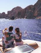 A couple relaxes on the deck of a yacht, embracing and pointing towards the scenic coastline of Los Cabos under a bright blue sky.