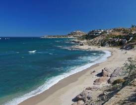 A high-angle, scenic view of Acapulquito Beach (also known as Old Man’s) in San Jose del Cabo. The image shows rolling azure waves perfect for longboarding, a pristine stretch of light sand, and white villas perched on the rugged, sun-drenched cliffs overlooking the Sea of Cortez under a deep blue sky.