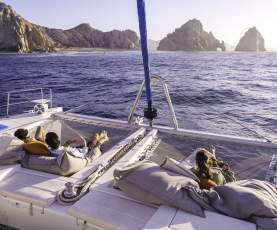 Passengers lounge on large cushions on the front deck of a luxury catamaran boat cruising toward Land's End and the Arch of Cabo San Lucas in Mexico.