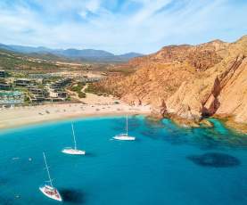 An aerial view of the vibrant turquoise waters of Santa Maria Bay in Los Cabos, featuring three white sailboats anchored in the calm, horseshoe-shaped cove. The golden sand beach is framed by dramatic reddish-brown cliffs and the modern architecture of a luxury resort under a bright blue sky with wispy white clouds.