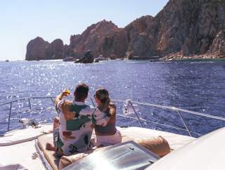 A couple relaxes on the deck of a yacht, embracing and pointing towards the scenic coastline of Los Cabos under a bright blue sky.