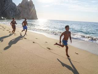 A family seated at an outdoor beachfront restaurant under large umbrellas, enjoying a relaxed meal with a view of the ocean and a cruise ship in the distance.