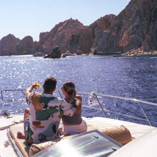 A couple relaxes on the deck of a yacht, embracing and pointing towards the scenic coastline of Los Cabos under a bright blue sky.