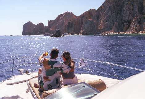 A couple relaxes on the deck of a yacht, embracing and pointing towards the scenic coastline of Los Cabos under a bright blue sky.