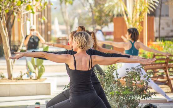 A group of women participate in an outdoor yoga class, holding the Warrior II pose with arms outstretched. The shot focuses from the side on a blonde woman in a black tank top and pants, with other participants and sunny resort landscaping softly blurred in the background.
