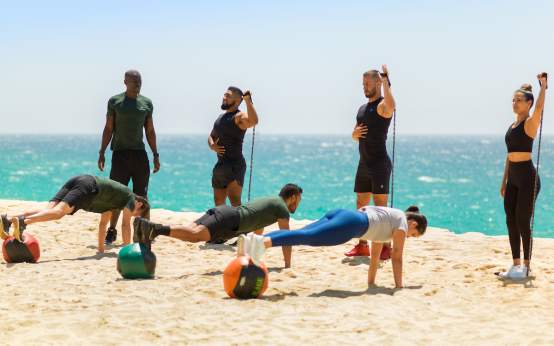 Alt Text:
An outdoor group fitness class takes place on a sandy beach with a bright turquoise ocean in the background. In the foreground, three participants hold plank positions with their feet elevated on colorful medicine balls. Behind them, other individuals exercise with resistance bands under a clear blue sky.