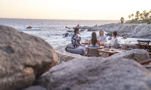 A group of people enjoying a meal outdoors with a scenic ocean view at sunset, framed by natural rocks.