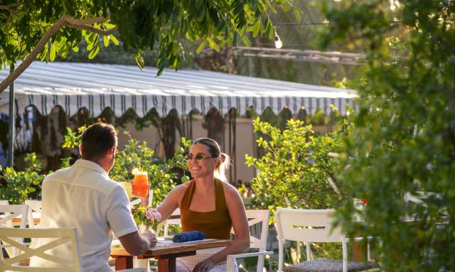 A couple enjoying an outdoor dining experience at a garden restaurant, surrounded by lush greenery, with the woman smiling and raising a glass of orange drink