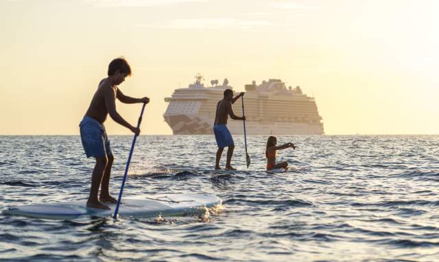 A group of people paddleboarding, including a child standing and others paddling, with a large cruise ship in the distance.