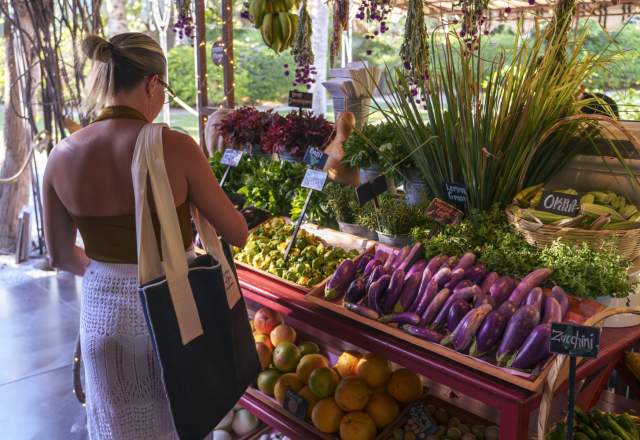 A woman shopping at a colorful market stall filled with fresh vegetables, herbs, and fruits, illuminated by soft sunlight filtering through the trees