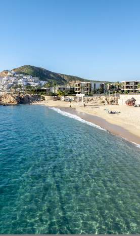 An aerial, high-angle view of the crystal-clear, turquoise waters and golden sands of Chileno Bay in Los Cabos. The swimmable beach is lined with modern luxury resorts and framed by rugged desert hills under a bright, clear blue sky.