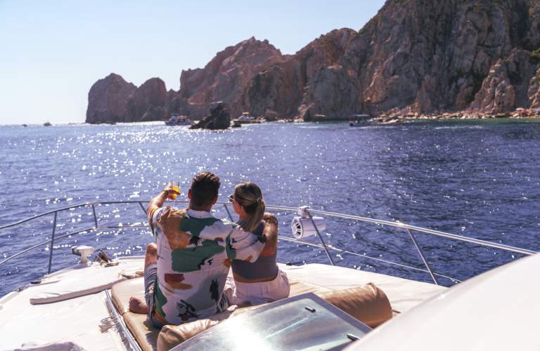 A couple relaxes on the deck of a yacht, embracing and pointing towards the scenic coastline of Los Cabos under a bright blue sky.