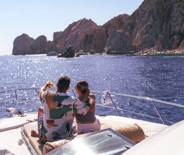 A couple relaxes on the deck of a yacht, embracing and pointing towards the scenic coastline of Los Cabos under a bright blue sky.