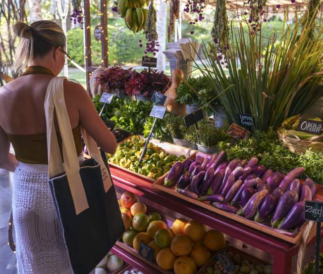 A woman shopping at a colorful market stall filled with fresh vegetables, herbs, and fruits, illuminated by soft sunlight filtering through the trees