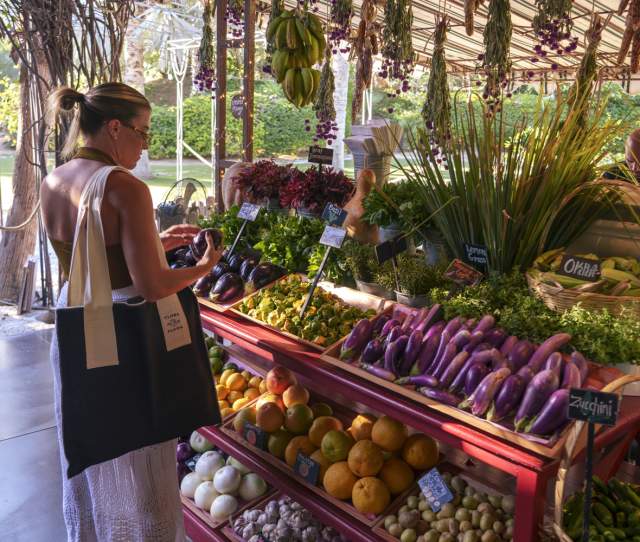 he same woman closely examining vegetables at the market stall, surrounded by vibrant produce, including eggplants, zucchini, and citrus fruits