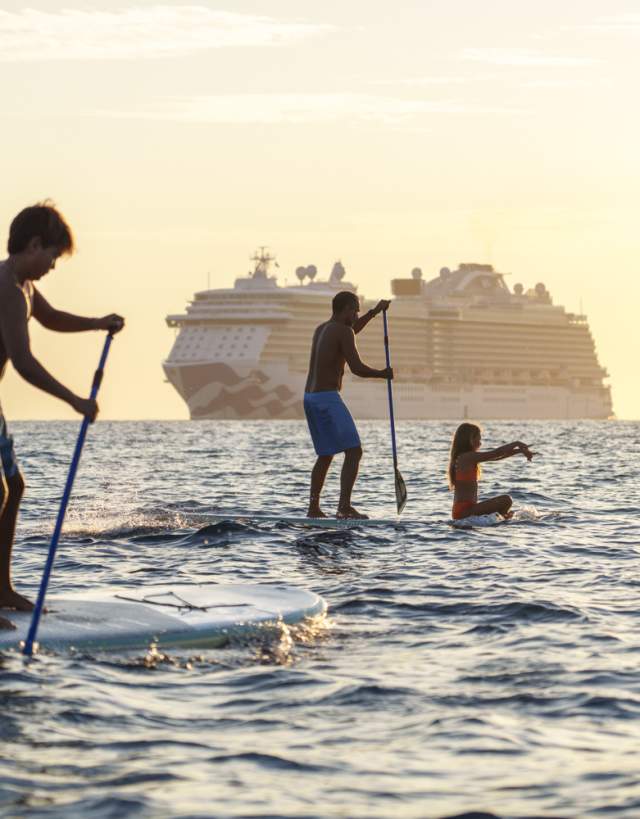 A group of people paddleboarding, including a child standing and others paddling, with a large cruise ship in the distance.