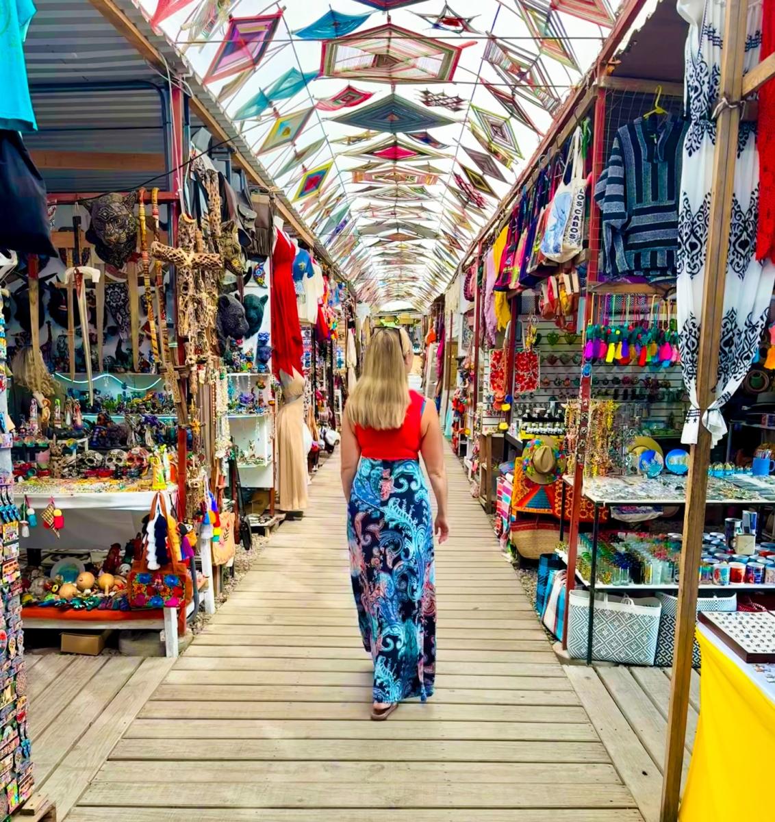 A woman in a red top and patterned blue skirt walks down a wooden path through a vibrant artisan market in Todos Santos. The ceiling is decorated with dozens of colorful "Ojos de Dios" (God’s Eyes) yarn crafts, while stalls on either side overflow with traditional Mexican textiles, masks, and handmade souvenirs.