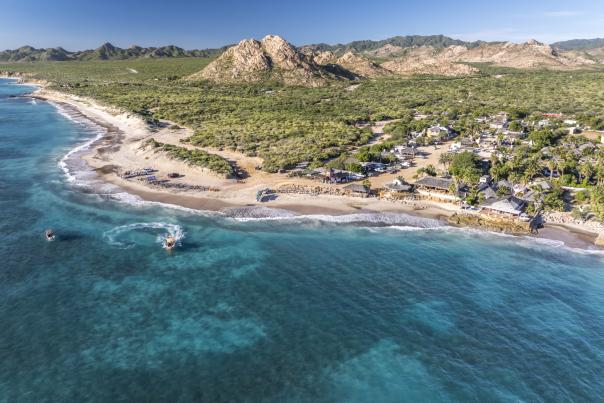 An aerial view of  Cabo Pulmo surrounded by lush greenery and rocky hills, with turquoise water and small boats near the shore