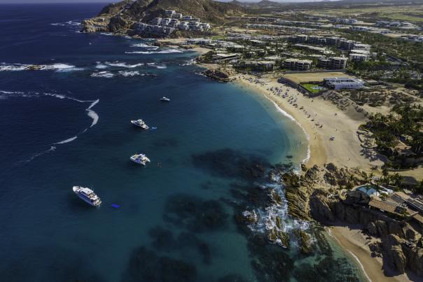 Aerial view of the crescent-shaped Chileno Bay in Los Cabos, featuring vibrant turquoise water, white yachts at anchor, and a golden sand beach lined with luxury resorts and rugged desert hills under a clear blue sky.