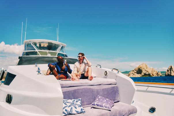 Two men relaxing on the cushioned bow of a luxury white yacht, holding champagne glasses with the ocean and the iconic rock formations of Cabo San Lucas in the background under a clear blue sky.