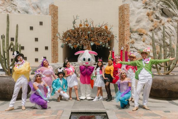 A group of performers in vibrant, whimsical Easter costumes posing with a large Easter Bunny mascot at the entrance of the Waldorf Astoria Los Cabos Pedregal. They are gathered in front of the iconic Dos Mares Tunnel, surrounded by desert rock formations and tall cacti in a festive, sunny scene.