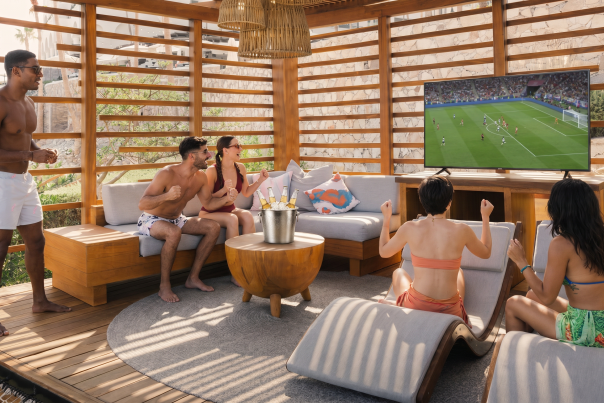 A group of friends in swimwear cheer while watching a soccer match on a large flat-screen TV inside a luxurious, open-air wooden cabana. An ice bucket filled with bottled beer sits on a round wooden coffee table in the center, surrounded by comfortable gray cushioned lounge seating in a sunny resort setting.