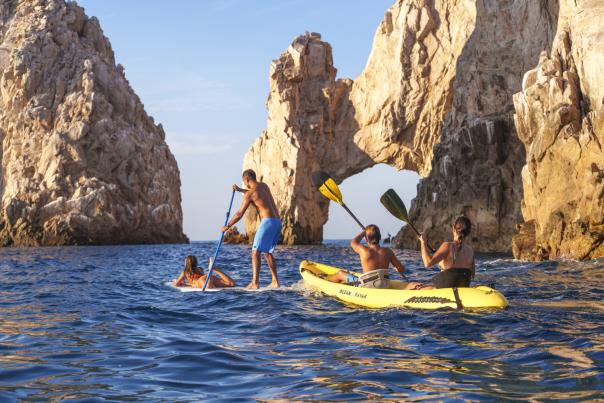A group enjoying water activities near "El Arco" in Los Cabos; one person on a stand-up paddleboard and two others kayaking in calm blue waters.