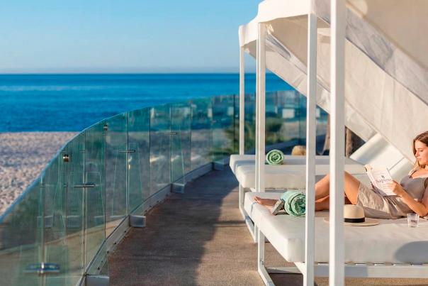 Mujer joven relajada en un camastro blanco, leyendo un libro. A su lado, sobre el camastro, hay una toalla de color turquesa, una botella de agua y un vaso. Al fondo se ve la arena de la playa y el mar azul de Los Cabos, bajo un cielo despejado. La escena tiene lugar en una terraza del Paradisus Los Cabos.