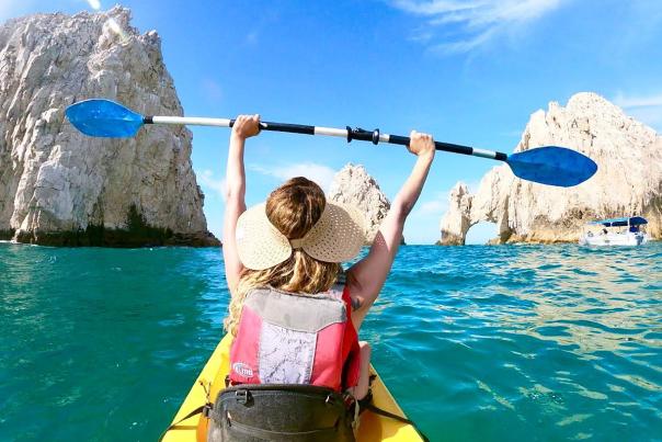 A traveler in a yellow kayak holds a blue paddle overhead in celebration while facing the iconic El Arco (The Arch) rock formation at Land's End in Cabo San Lucas, surrounded by vibrant turquoise water and rugged desert cliffs.