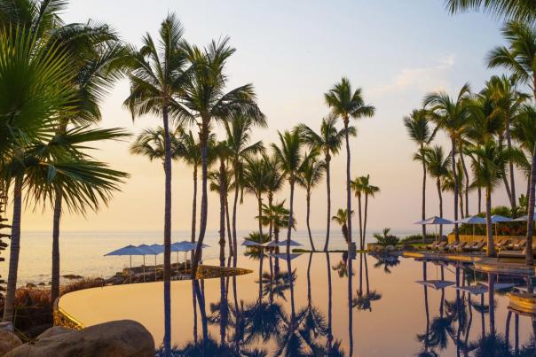 Tall palm trees are reflected in the still, glassy water of an infinity pool at the One&Only Palmilla resort in San José del Cabo, Mexico, during a calm sunrise over the ocean.
