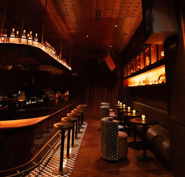 Dimly lit upscale cocktail lounge featuring a long backlit bar with leather barstools, black-and-white checkered flooring, and small table seating with ambient lamps.
